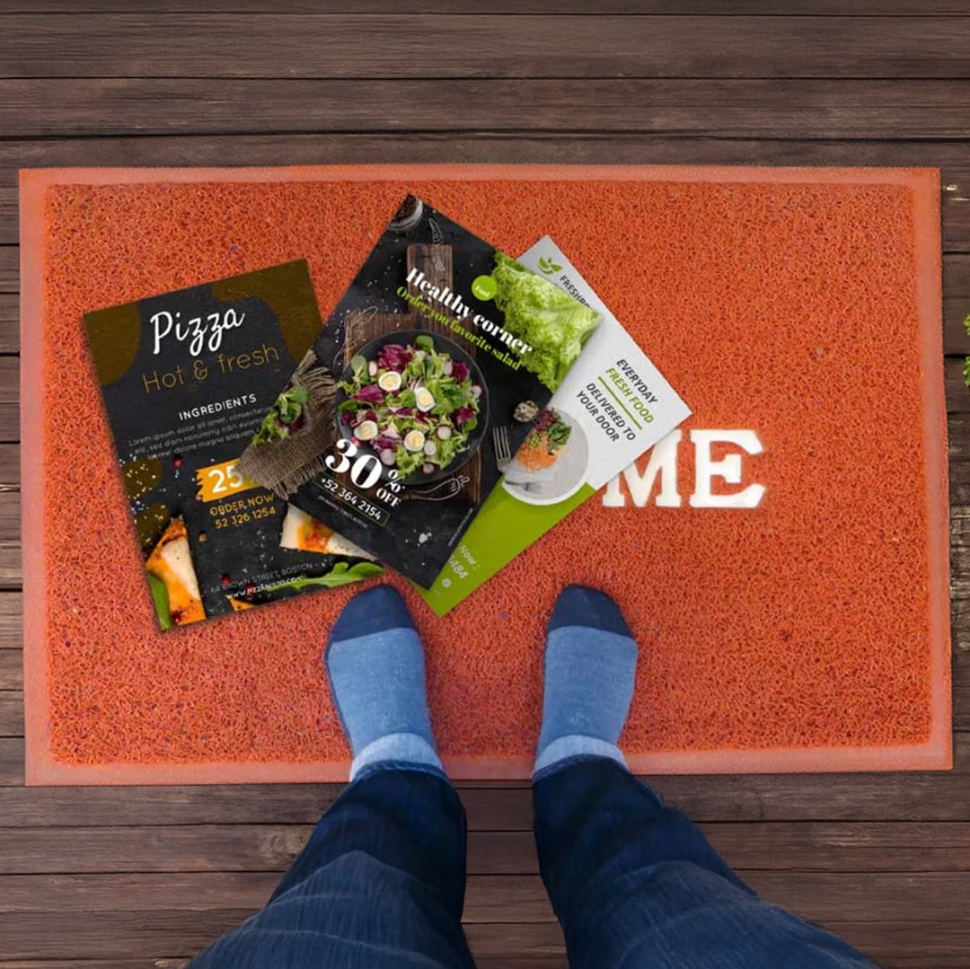 A person standing on a doormat, with a selection of leaflets on top.