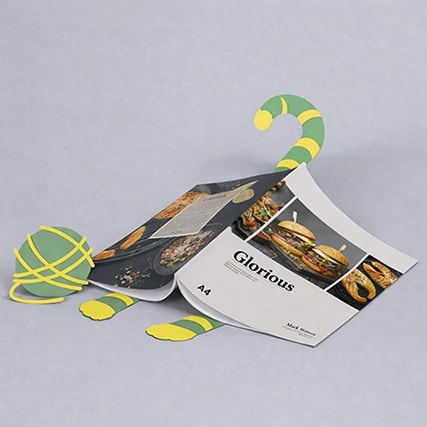 A magazine is propped open by a flat, cartoon-like lizard cutout on a light gray background.