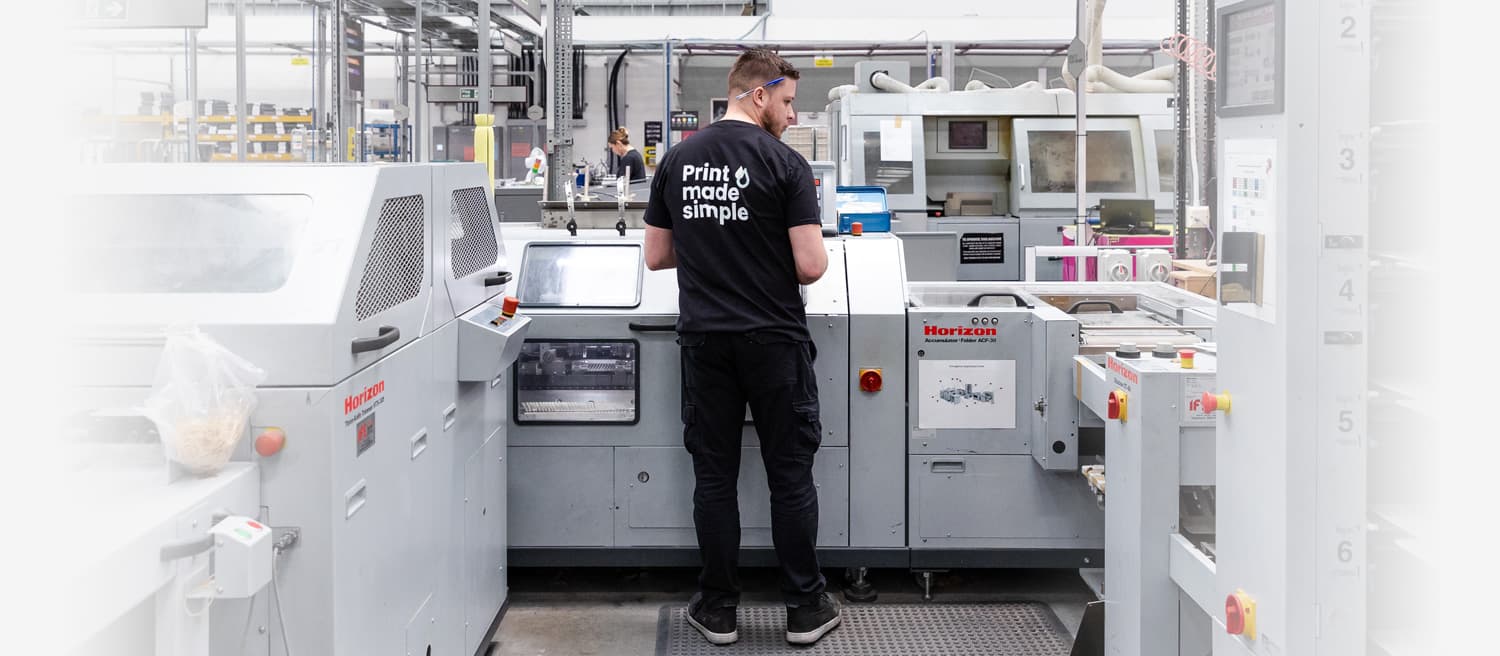 Person standing and operating a large printing machine in a factory. The back of their shirt reads, "Print made simple.