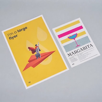 Two A3 posters on a gray surface; one yellow with a paper plane, one colorful with a cocktail.