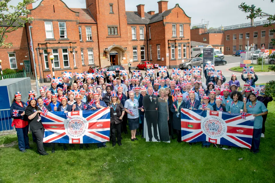 NHS workers gather with British flags and printed items in front of a building to commemorate King Charles III's coronation.