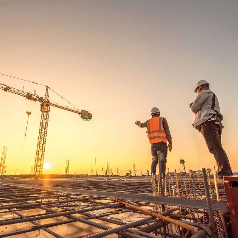 Two construction workers stand on a building site at sunset with a crane in the background.
