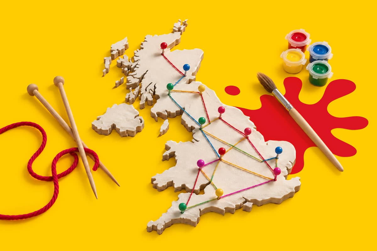 Wooden UK map with colored pins and string, knitting needles, yarn, paint, and brush on yellow.