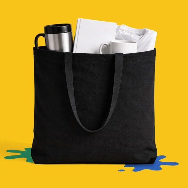 A black tote bag holding a tumbler, book, mug, and folded shirt, on a yellow background.