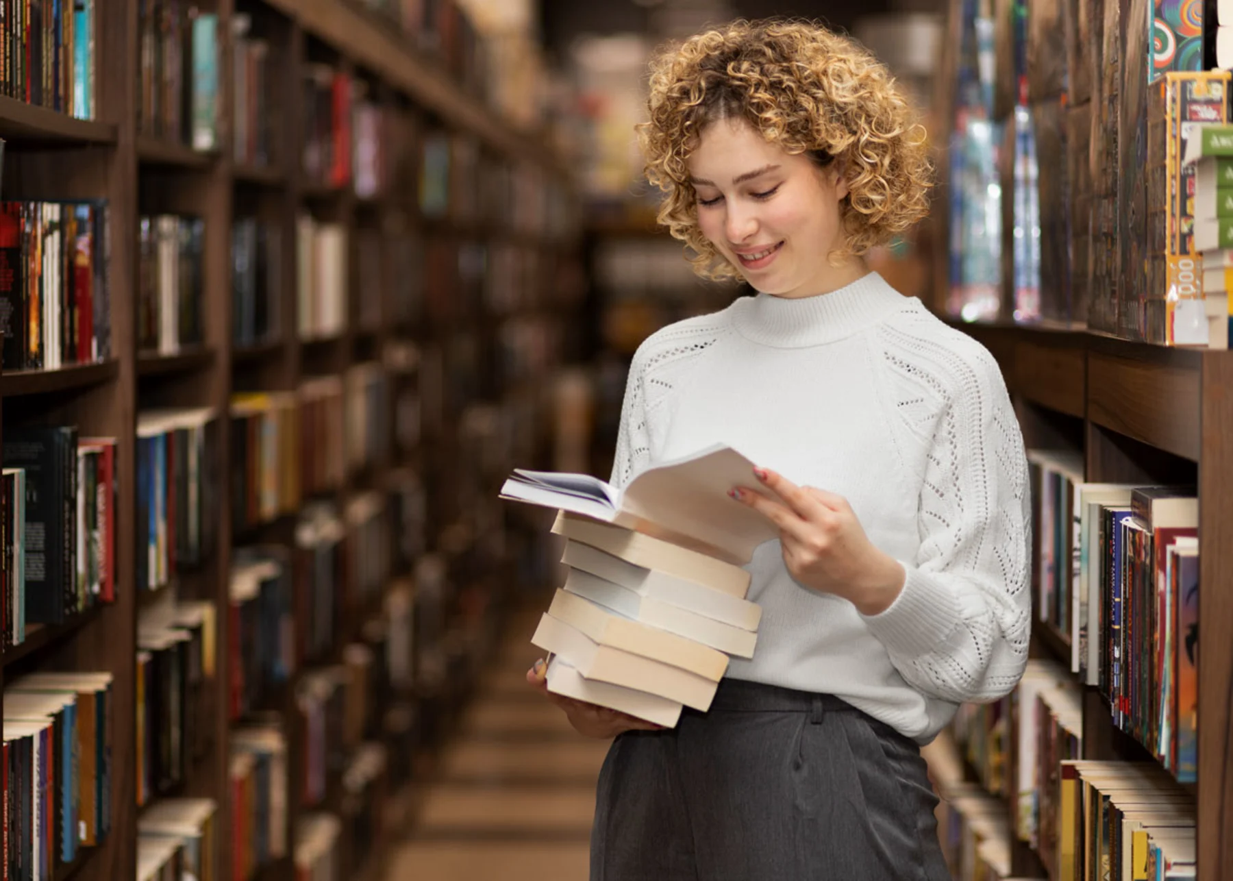 A woman stands in a library, holding a book, surrounded by shelves filled with various books