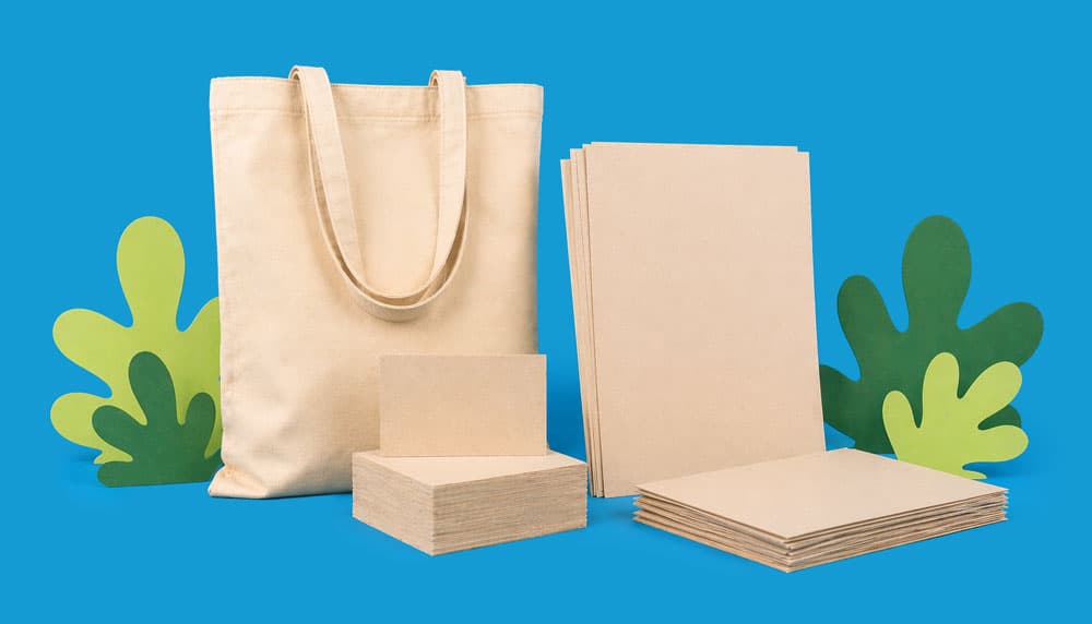 A canvas tote bag and stacks of sustainable paper against a blue background with green cutouts.