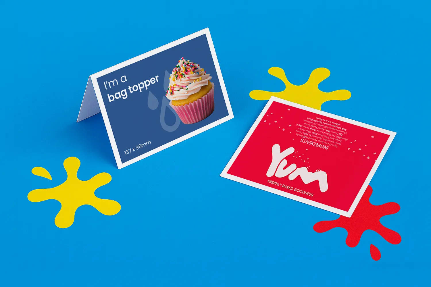 Two colourful printed bag topper cards on a blue surface with yellow and red paint splatters.