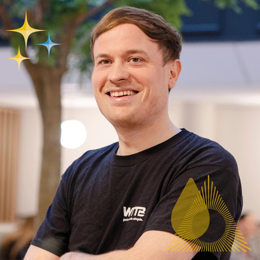 Smiling person wearing a black "WTB" T-shirt, standing indoors with a blurred background.