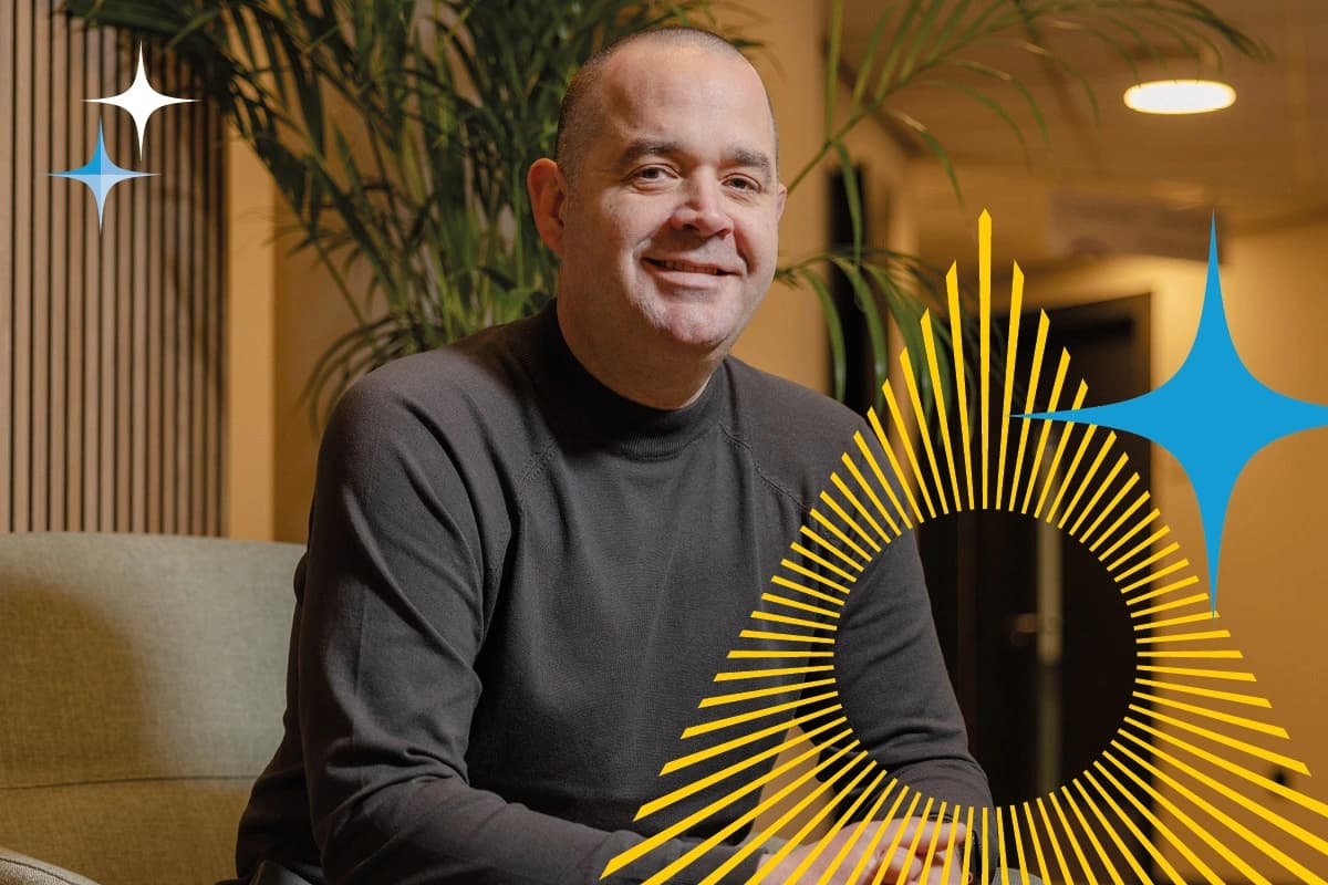 A man in a dark sweater sits indoors with graphic shapes and plants in the background.