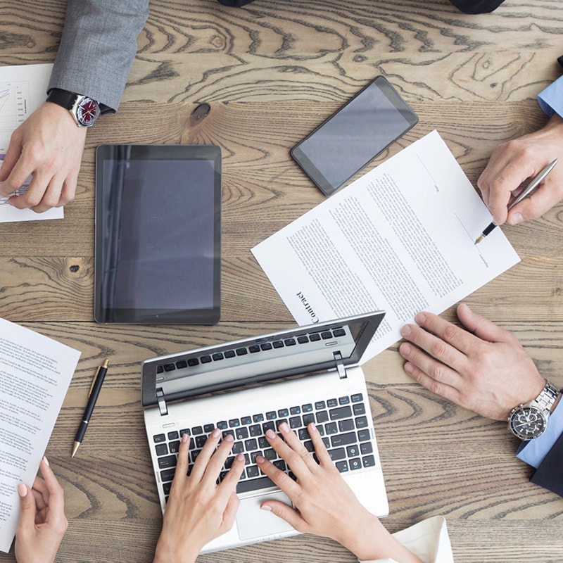 People working at a table with a laptop, documents, a tablet, and a smartphone.