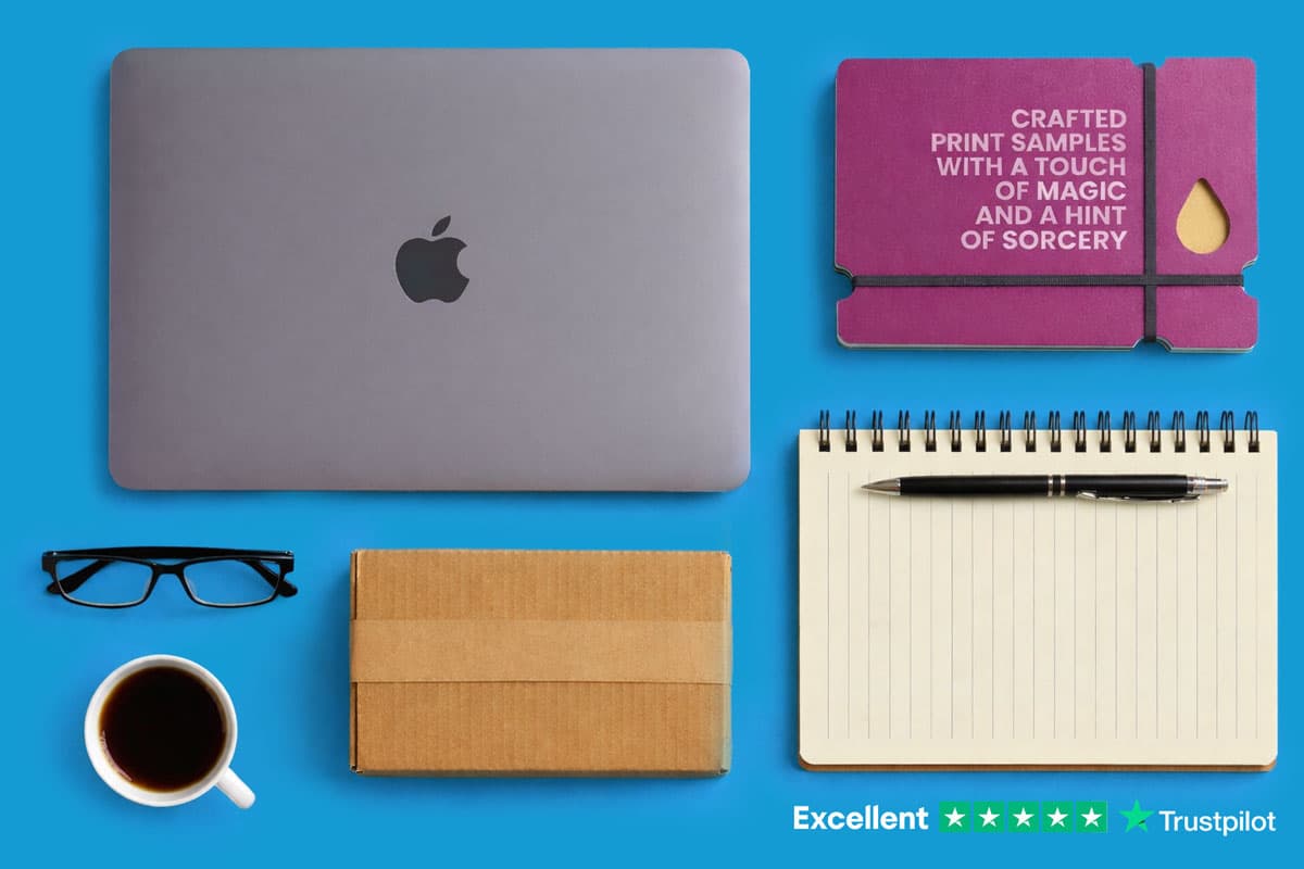 Laptop, notebook with pen, glasses, coffee, box, and purple print samples on blue background.