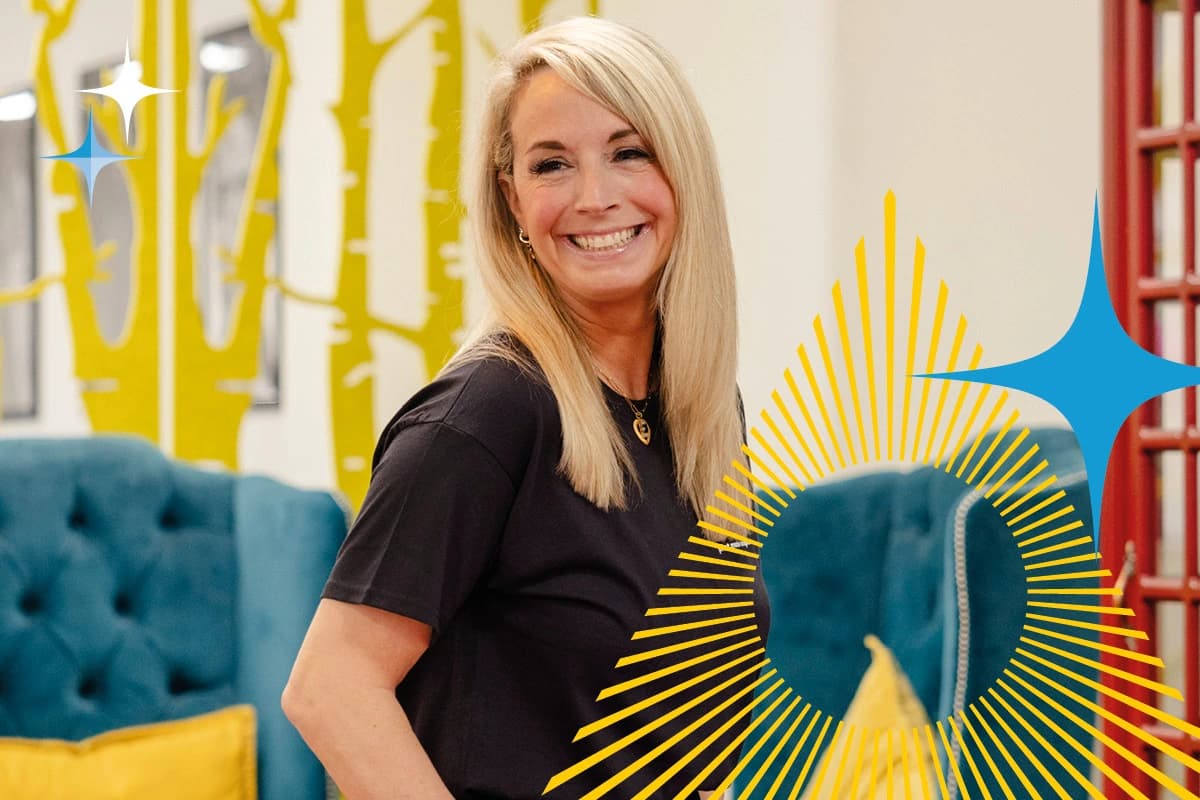 Woman with long blonde hair smiling, standing in a colourful modern room with yellow accents.