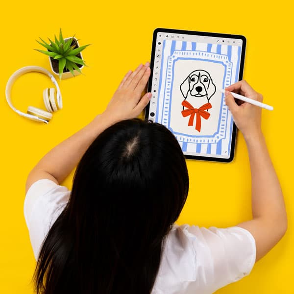 Person drawing a dog with a red bow on a tablet, with headphones and a plant beside them.