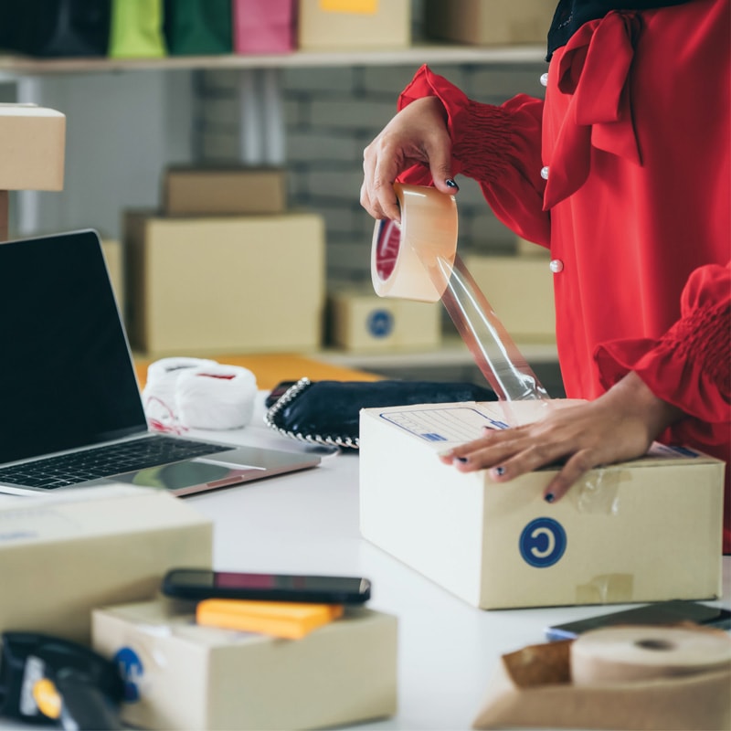 Person sealing a cardboard box with tape near a laptop and packages on a desk.