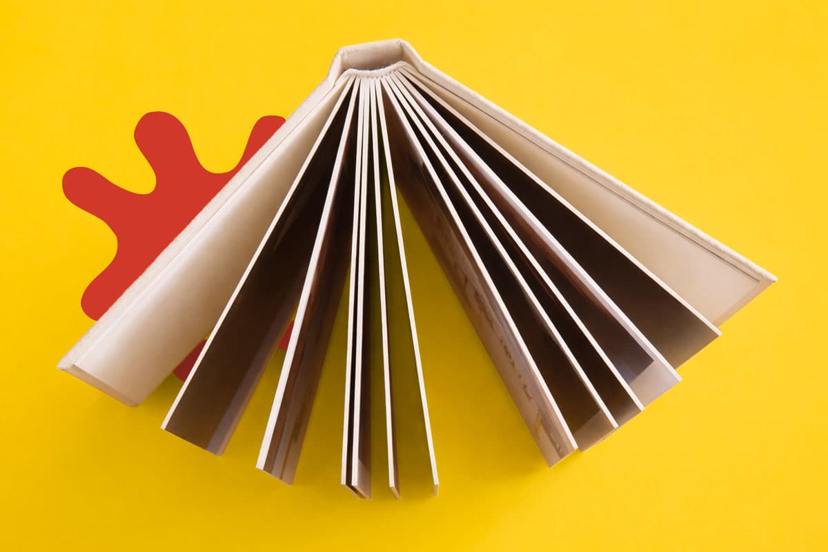 An open book fanned out on a yellow background with a red paint splatter shape.