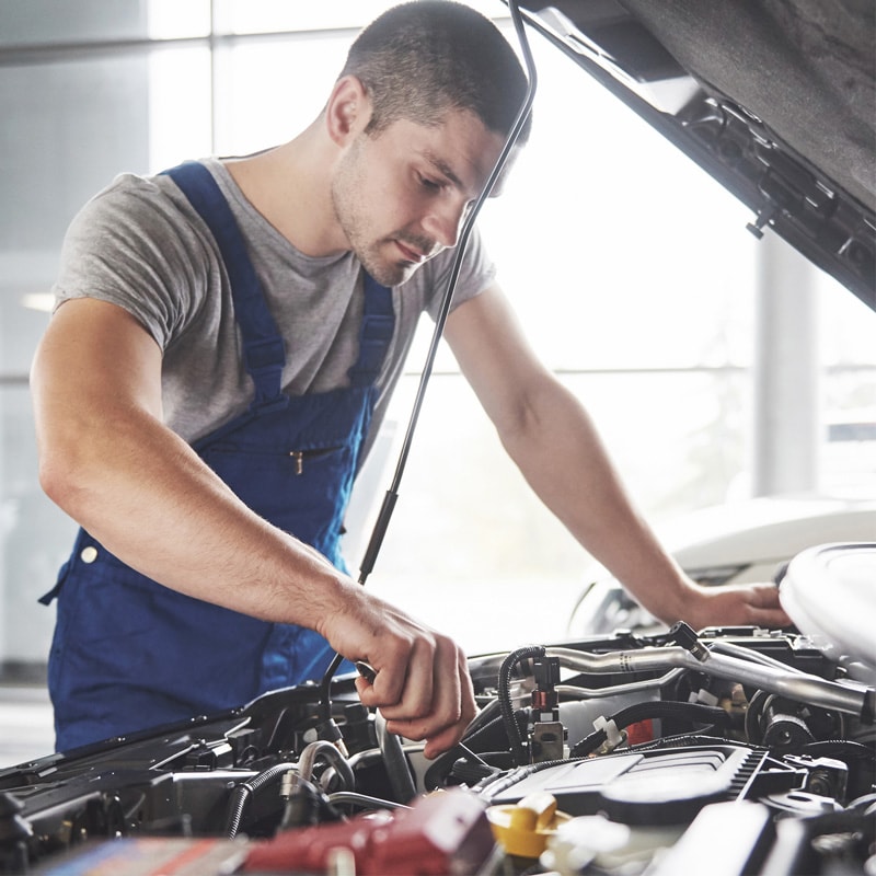 Mechanic in blue overalls working on a car engine with the hood open.