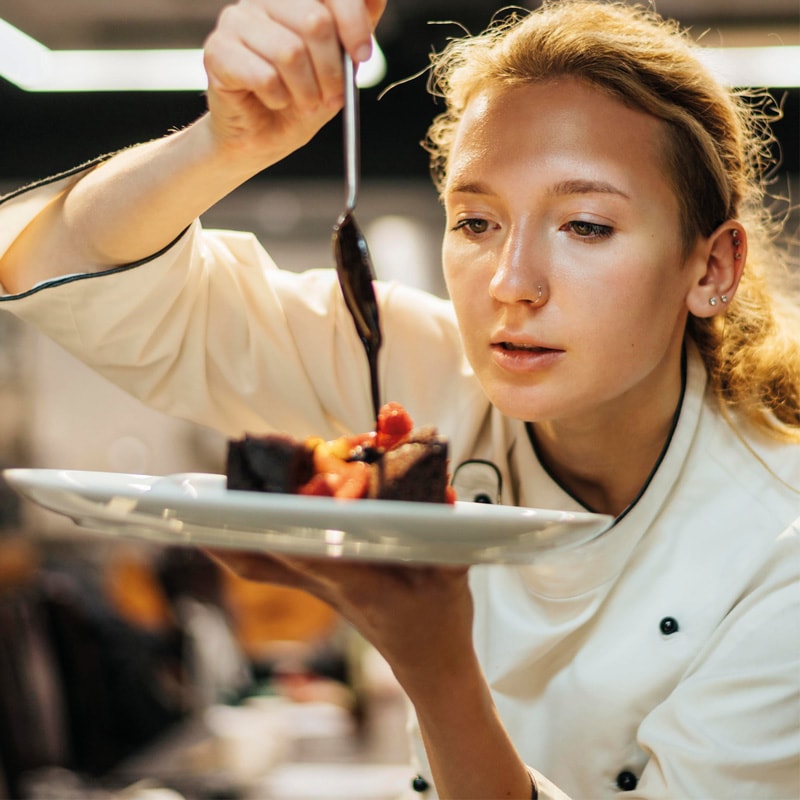 Chef garnishes a dessert with sauce on a plate in a professional kitchen.