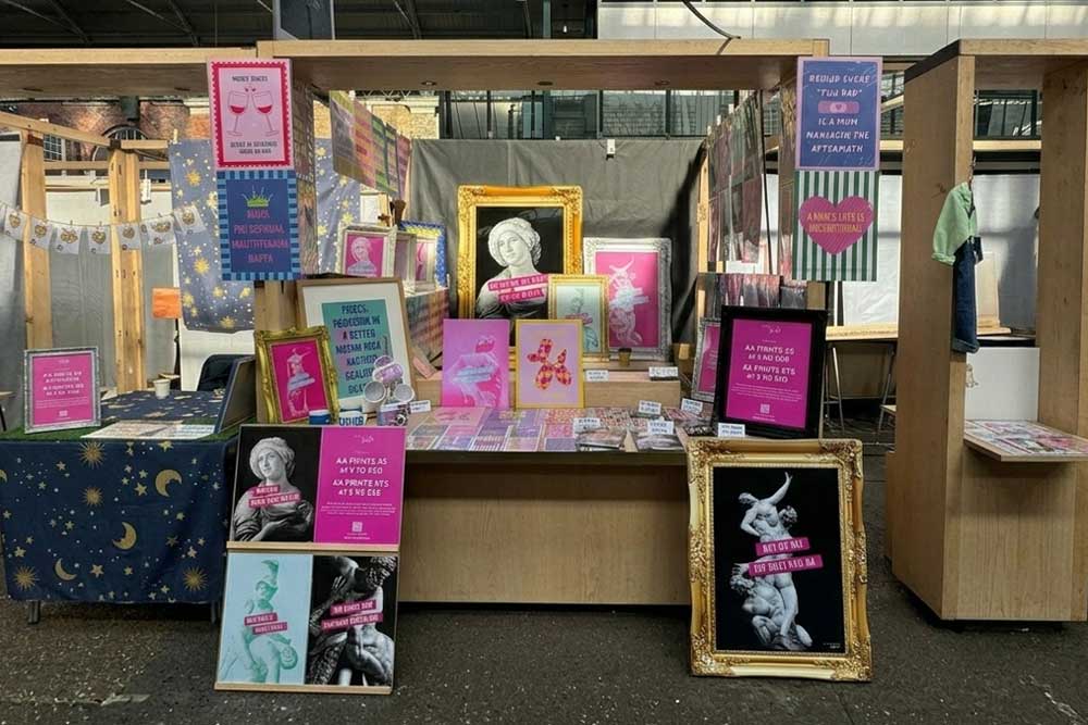 Art market stall displaying colourful framed prints and posters with humorous text, indoors.