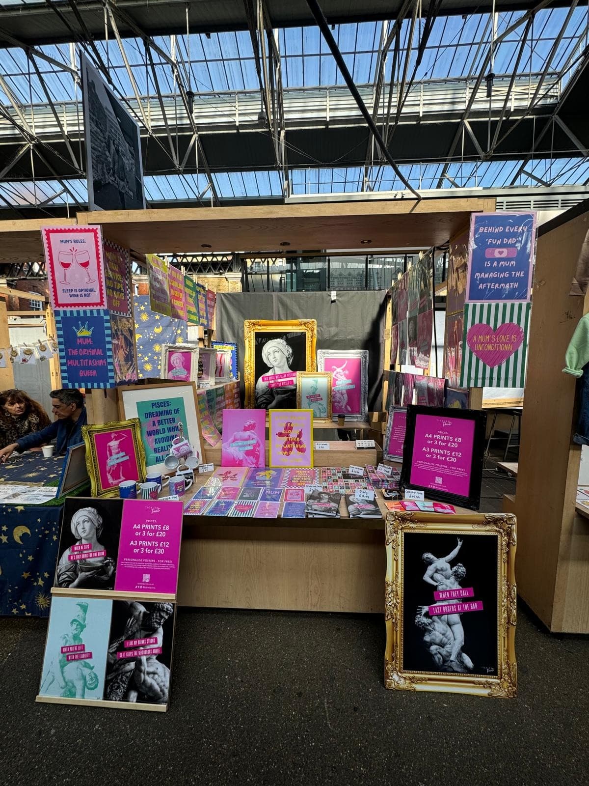 Art market stall displaying colourful framed prints and posters with humorous text, indoors.