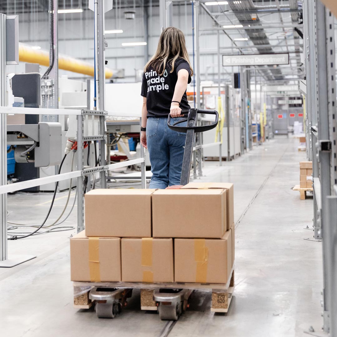 A woman navigates a cart loaded with boxes, operating a pallet truck while sporting a "Print Made Simple" t-shirt