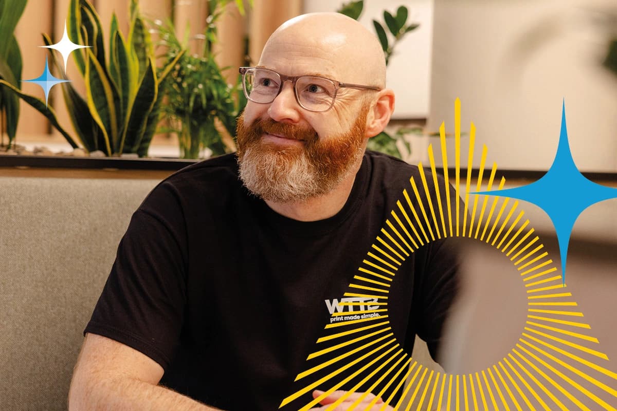 Bald man with glasses and beard sits at a table, smiling, with graphic elements overlaid.