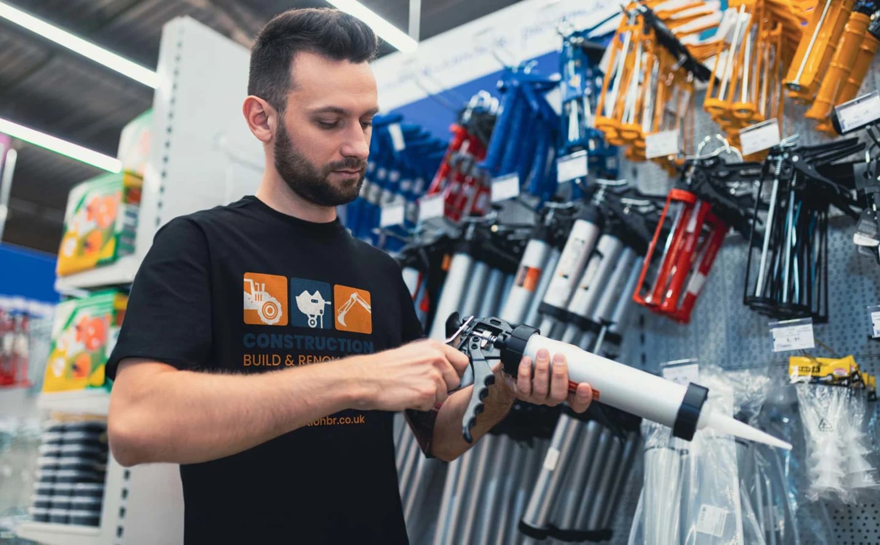 A man in a modern workwear t-shirt holds a hot glue gun in a hardware store, ready for a project