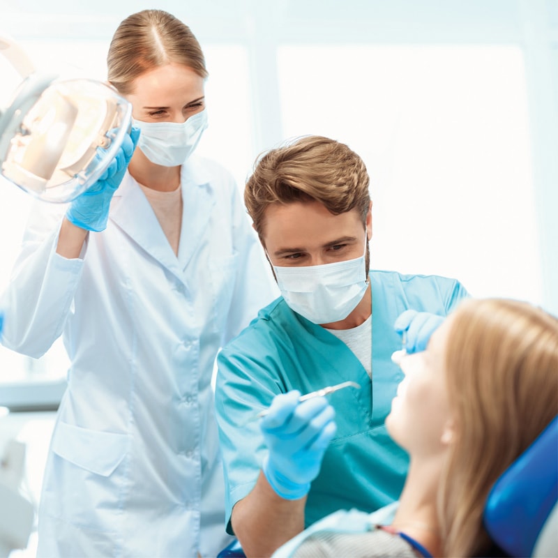 Two dentists examine a patient’s teeth in a dental clinic, wearing masks and gloves.