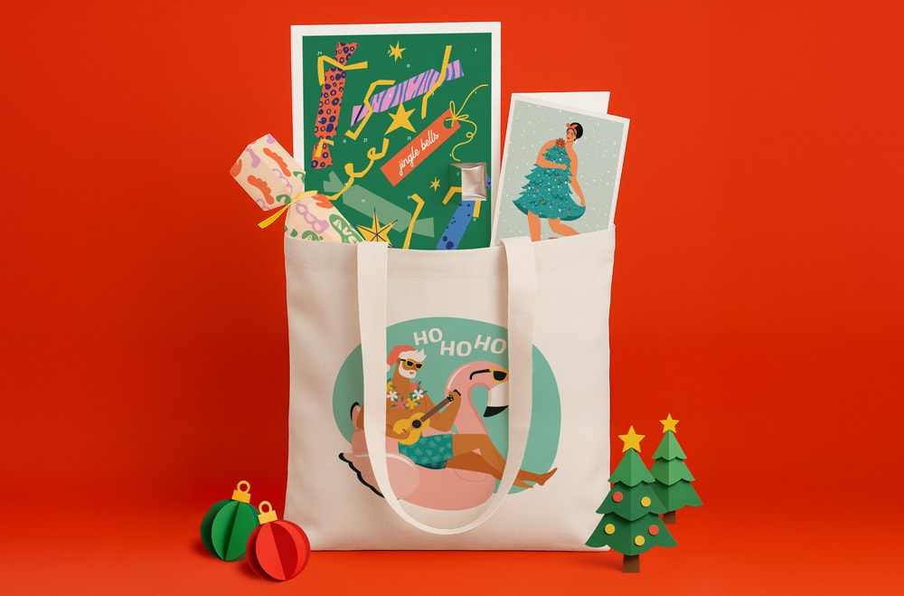 White tote bag with "Meowy Christmas" design filled with gifts, surrounded by ornaments and a tree.