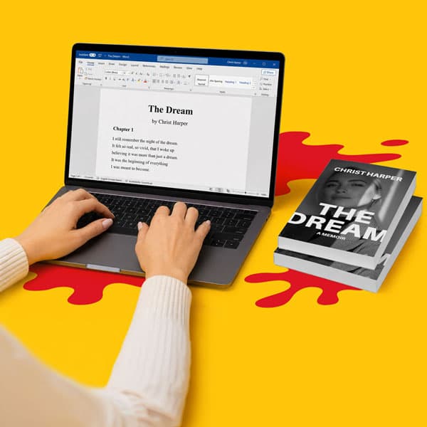Hands typing on a laptop with a manuscript and two copies of “The Dream” book on a yellow table.