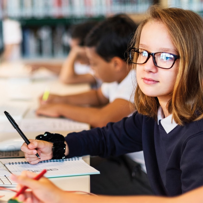 Student wearing glasses writing in a notebook at a classroom desk with others in the background.
