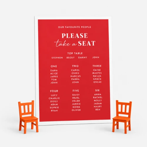 Red wedding seating chart sign with names, flanked by two small orange chairs.
