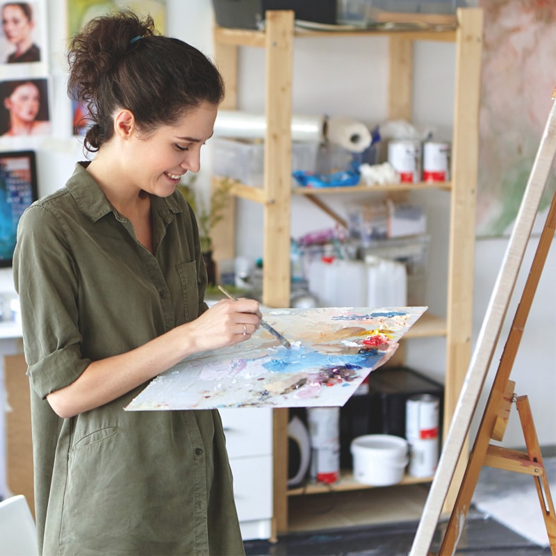 Woman painting on a canvas in an art studio, holding a palette and smiling.