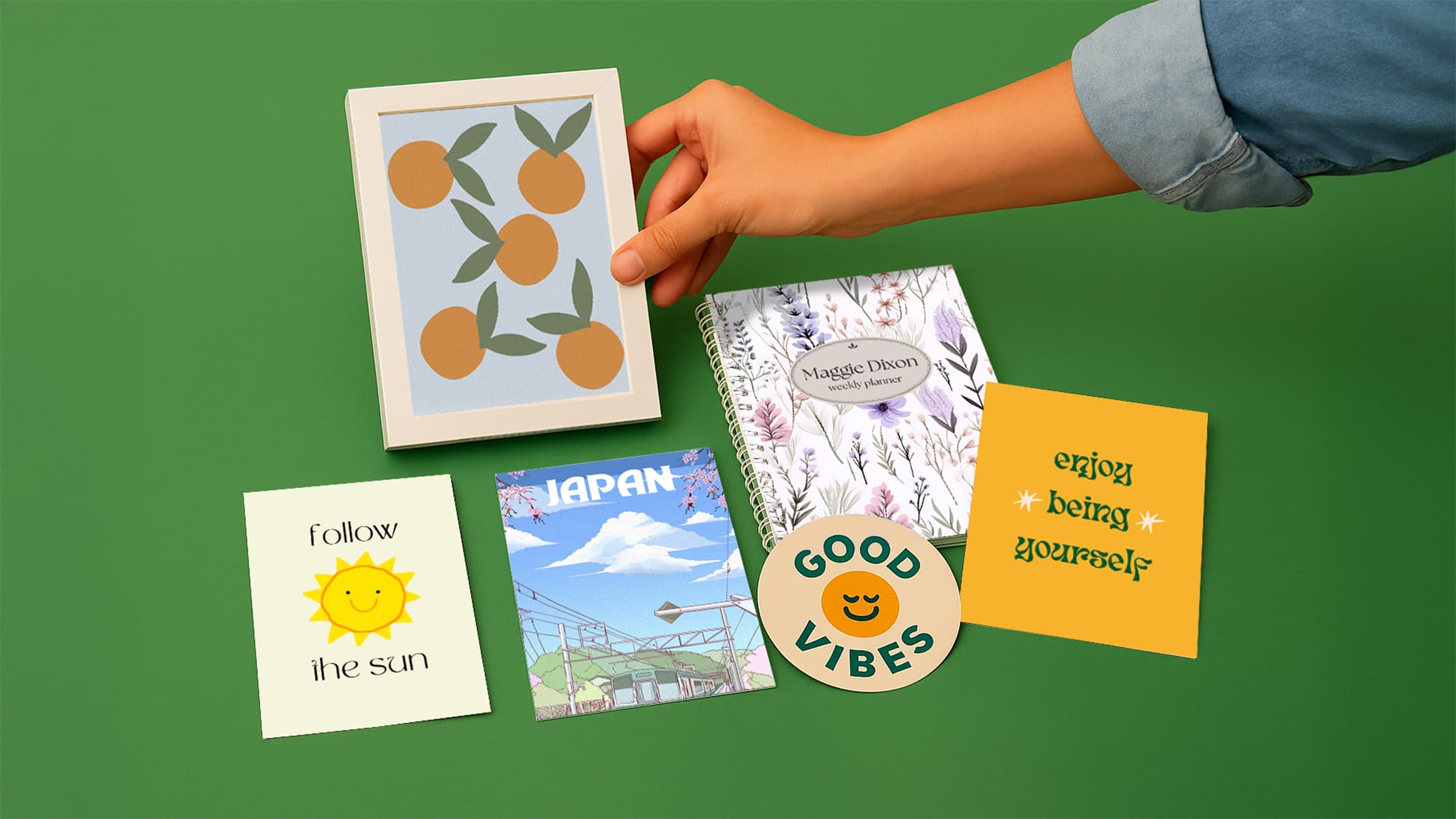 A hand arranges art prints, a notebook, and stickers on a green background.