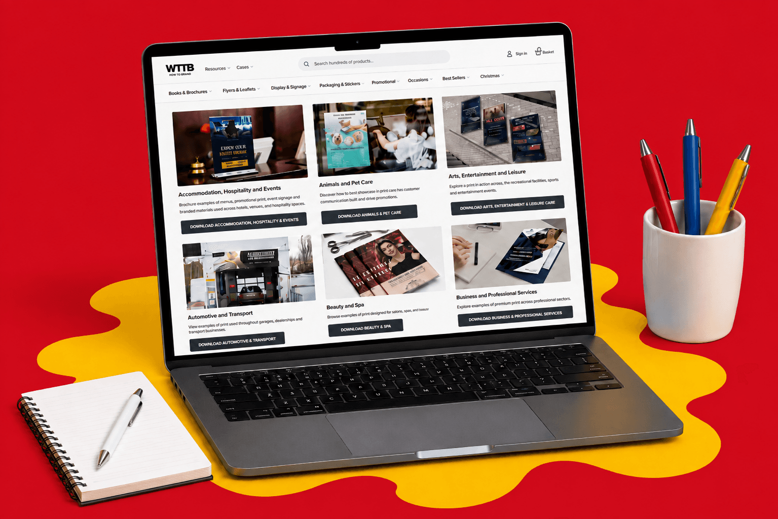 Laptop displaying an online image library; notebook and pens on desk with a red and yellow background.