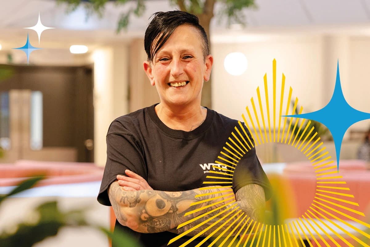 Person with short dark hair and tattoos smiles with arms crossed in a modern indoor setting.