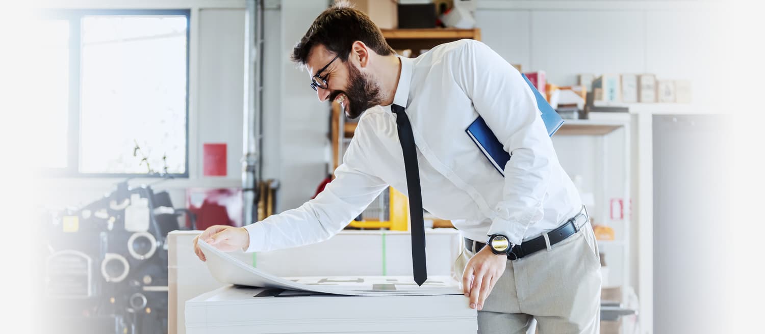 A man in a white shirt and tie stands before a printer, examining a stack of freshly printed sheets.