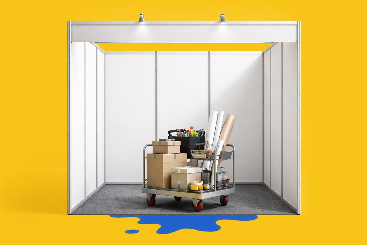 A metal cart with boxes and supplies sits inside an empty exhibition booth with a yellow background.