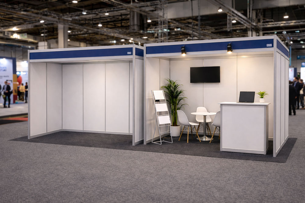 Two adjacent trade show booths; one empty, the other furnished with chairs, desk, and monitor.