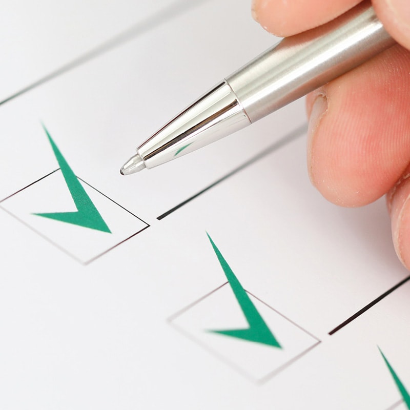 A hand holding a pen near checkboxes with green check marks on a checklist.