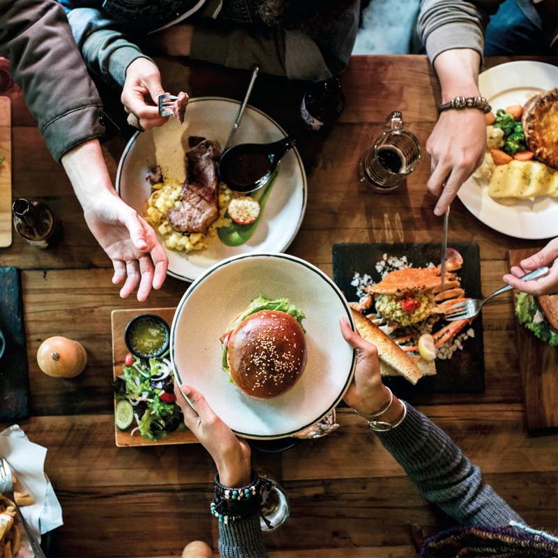 People sharing a meal with burgers, steak, salad, and fries on a wooden table.