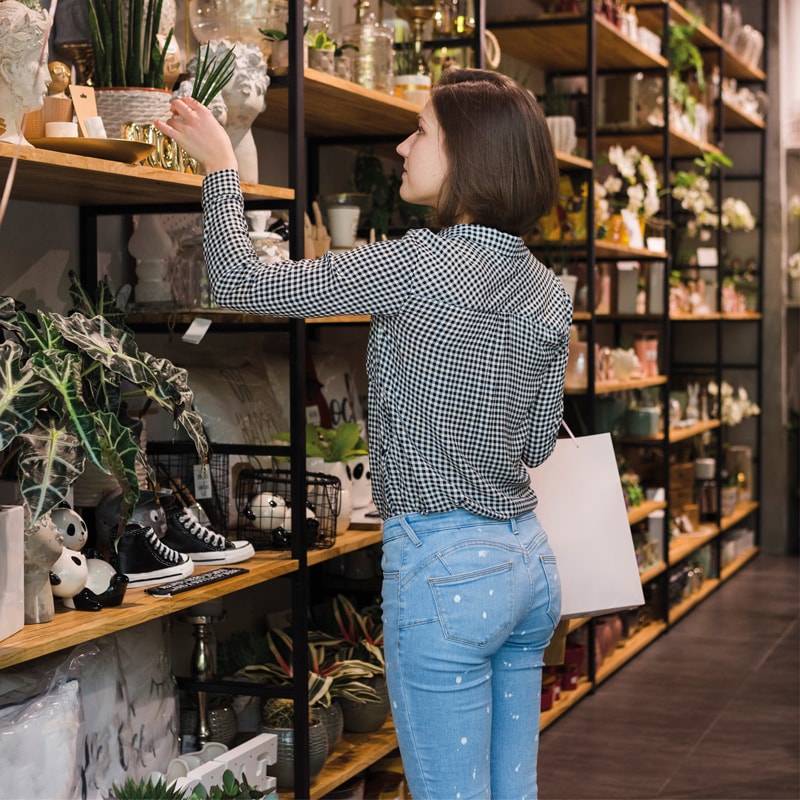 Woman browsing potted plants and decor items on shelves in a store, holding a shopping bag.