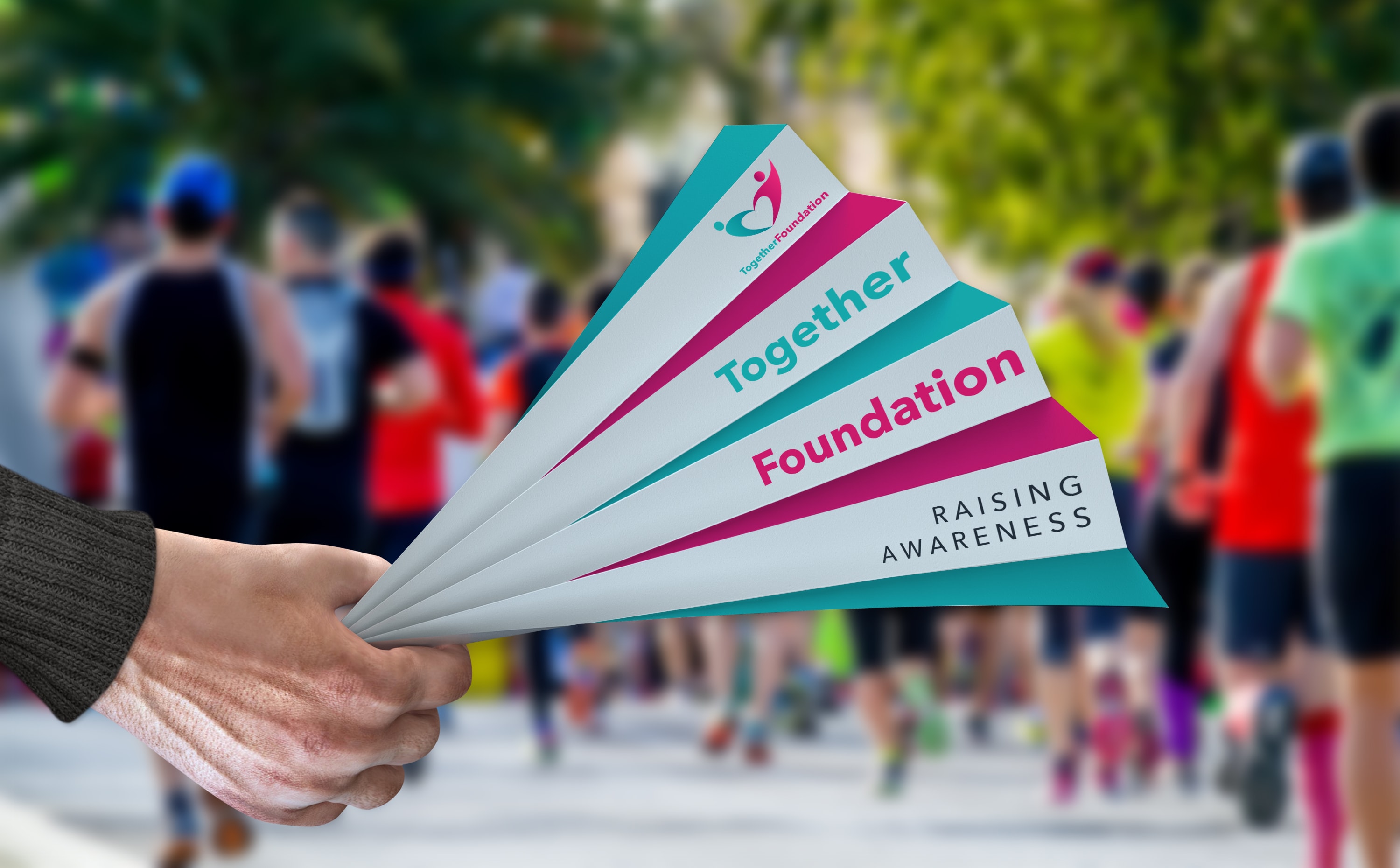 Hand holding promotional paper fan clappers at an outdoor charity run event, runners in background.