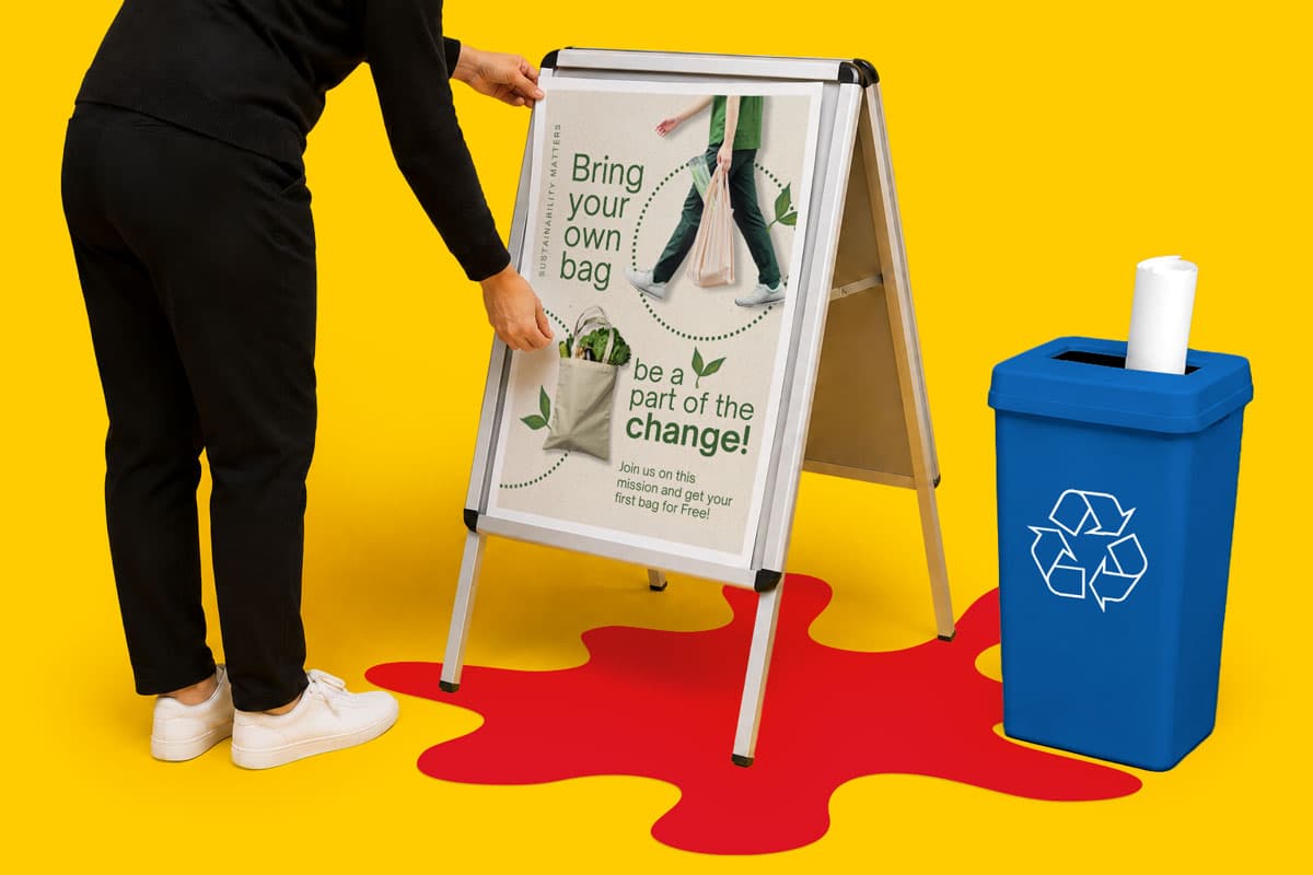 Person placing a "Bring your own bag" sign on an easel next to a blue recycling bin.