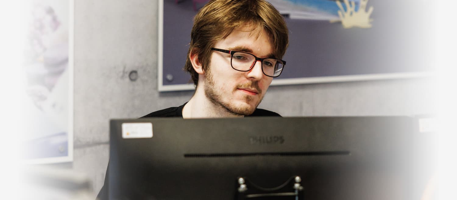 A man wearing glasses is seated at a computer, focused on the screen in a well-lit workspace.