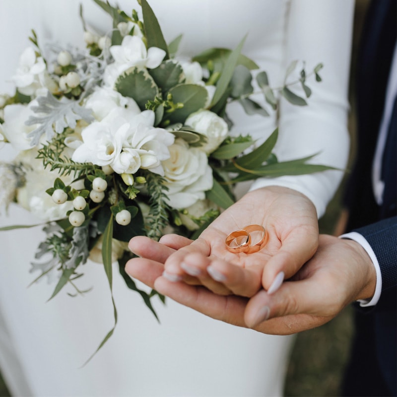 Two hands hold wedding rings; bouquet of white flowers and greenery in the background.