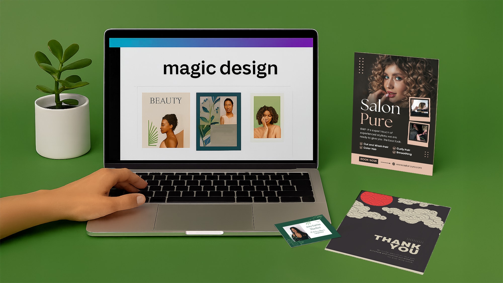 A hand uses a laptop displaying Canva design work, with printed materials and a plant nearby.