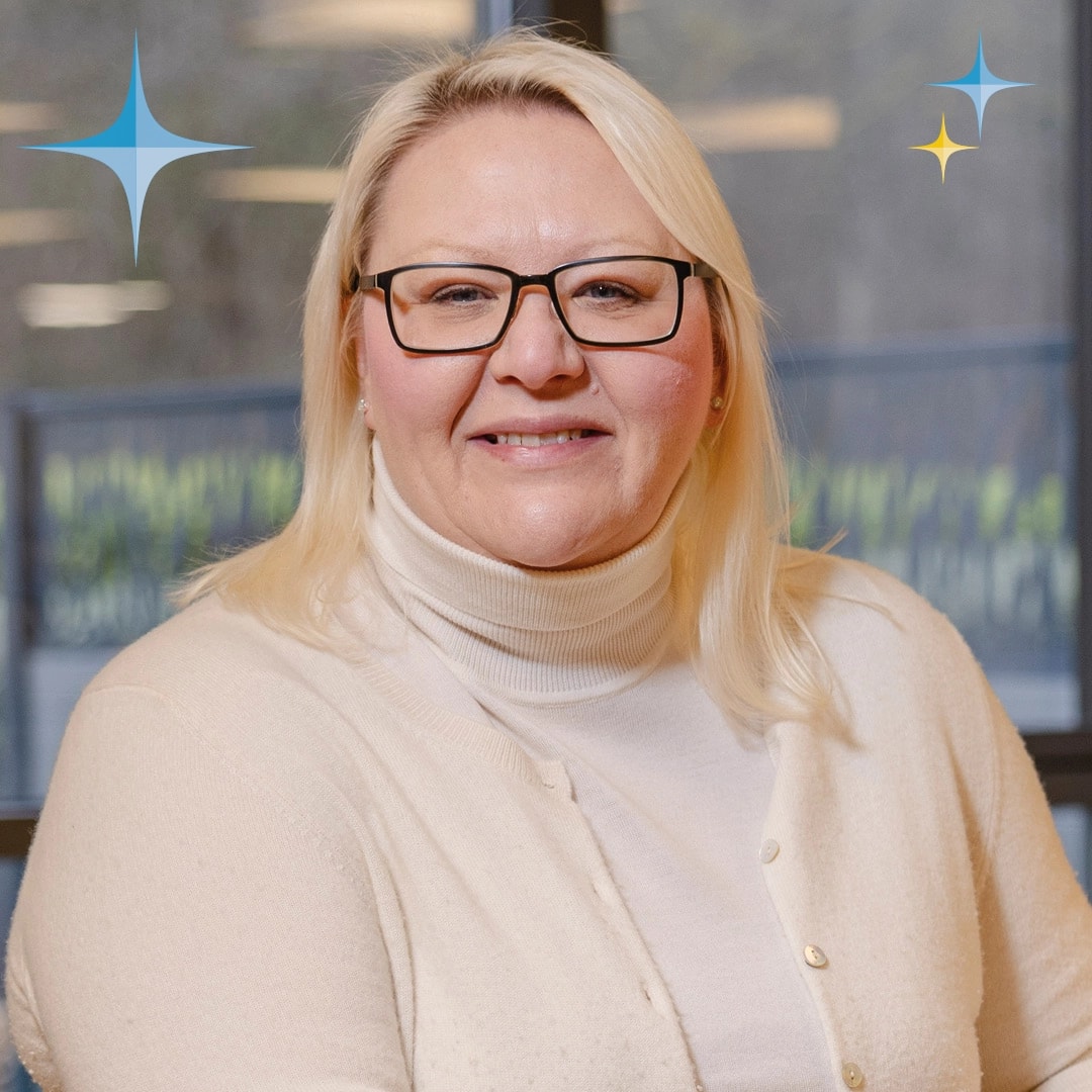 Woman with glasses and blonde hair smiling, wearing a white sweater, sitting indoors.