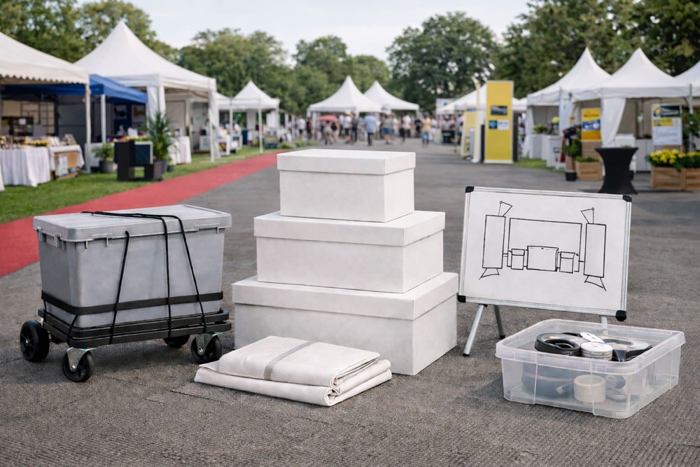 A gray cart, white boxes, folded cloth, a whiteboard, and a clear bin are on pavement.