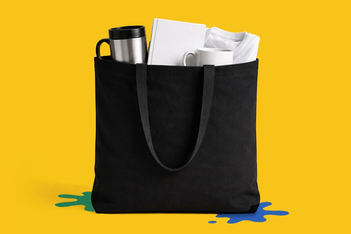 A black tote bag holding a mug, tumbler, t-shirt, and book on a yellow background.