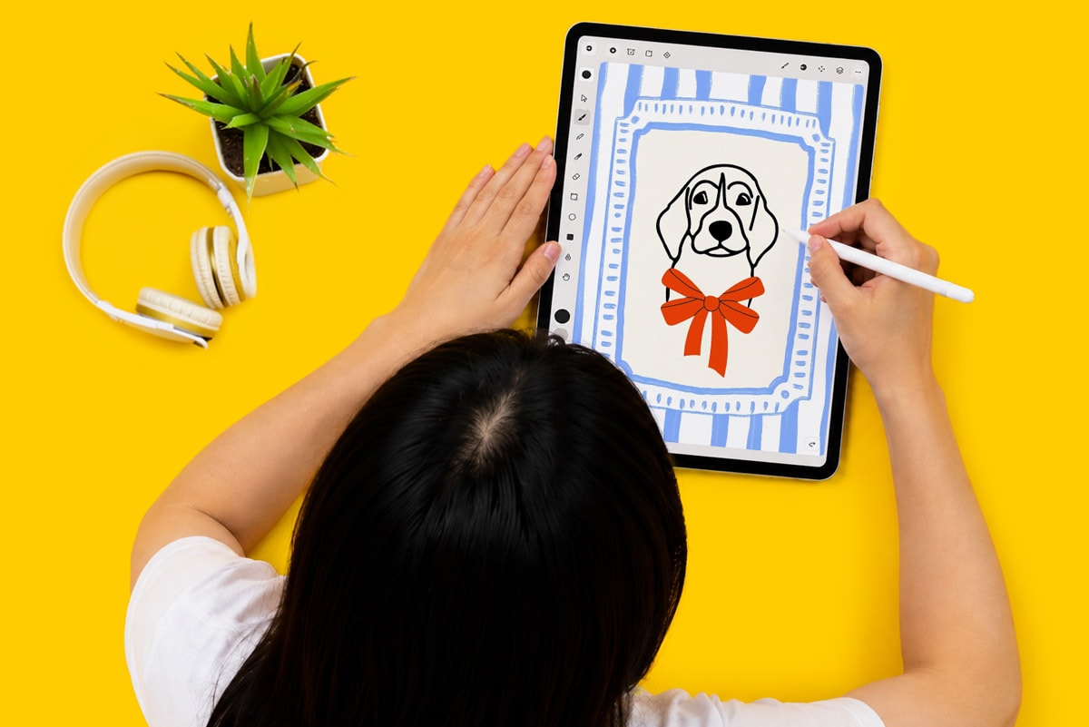 Person drawing a dog with a red bow on a tablet against a yellow background.